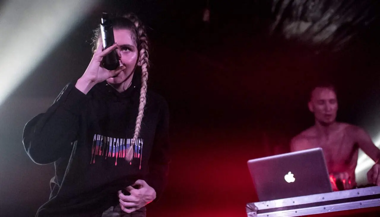 A person with long braids holds a microphone while performing on stage, wearing a black hoodie. In the background, another person operates a laptop with an Apple logo. The scene is lit with dramatic red and white stage lights.