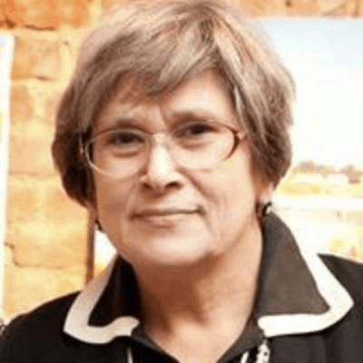 An older person with short gray hair, glasses, and a black collared shirt with white trim, smiling slightly in front of a blurred indoor background with a brick wall.