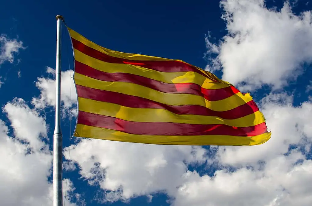 A yellow flag with four horizontal red stripes waves on a flagpole against a blue sky with scattered white clouds.