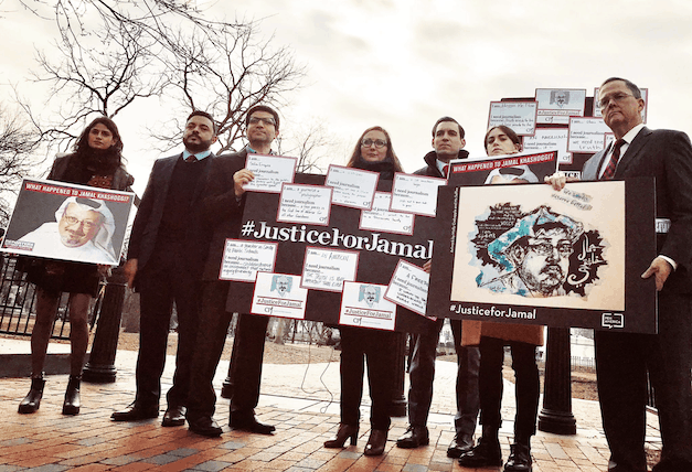 A group of people stand outdoors holding signs and posters calling for justice for Jamal Khashoggi. The signs feature hashtags, questions, and artwork related to Khashoggi’s case. Trees and a fence are visible in the background.