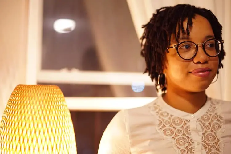 A woman with short dreadlocks and glasses stands indoors, wearing a white lace top. She is beside a woven lamp, with a softly lit window in the background.