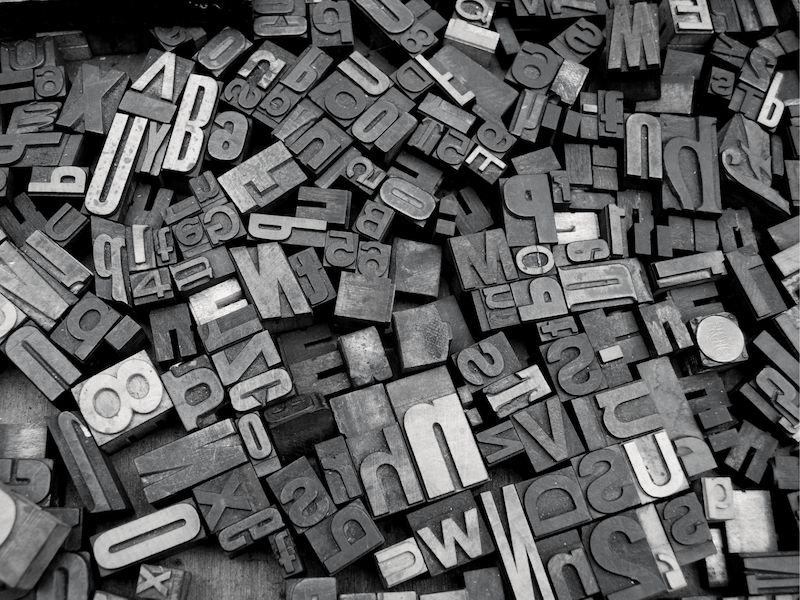A close-up of scattered vintage metal letterpress type blocks in various fonts and sizes, creating a chaotic, textured pattern in black and white.