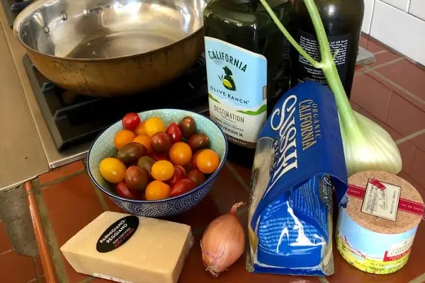 A variety of ingredients on a kitchen counter: a bowl of colorful cherry tomatoes, a wedge of parmesan cheese, a shallot, a fennel bulb, a package of spaghetti, sea salt, olive oil, and a sauté pan in the background.