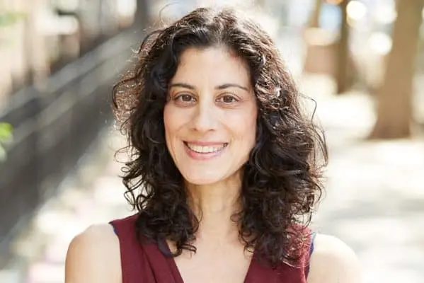 A woman with curly dark hair and a sleeveless maroon top smiles outdoors on a sunny day, with trees and a blurred sidewalk in the background.