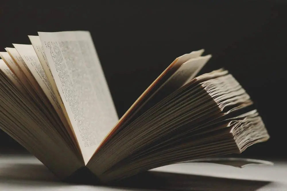 An open book sits on a flat surface with its pages slightly fanned out, illuminated by soft light against a dark background.