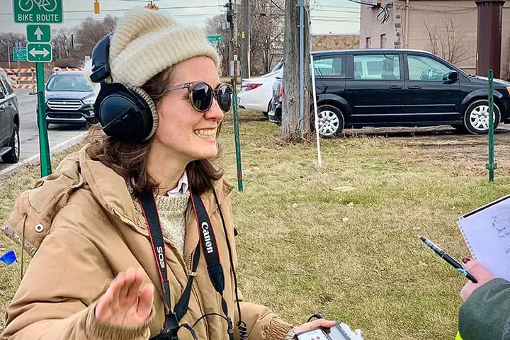 A smiling woman wearing a beanie, sunglasses, and headphones stands outside on a grassy area. She has a camera around her neck and gestures with her hand, while holding a notepad. Cars and street signs are visible in the background.