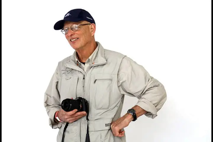 An older man wearing glasses, a navy baseball cap, and a light beige jacket smiles while holding a camera, standing against a plain white background.