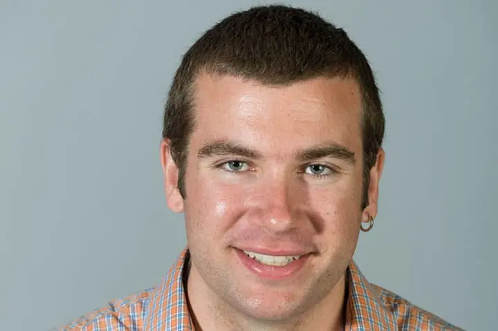 A young man with short brown hair, wearing a plaid shirt and a silver hoop earring, smiles at the camera against a plain gray background.