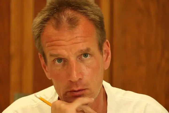 A man with short brown hair holds a pencil and looks thoughtfully at the camera, resting his chin on his hand. He is wearing a white shirt and sits in front of a wooden background.
