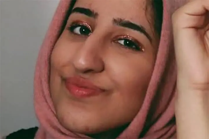 A person wearing a pink hijab smiles softly at the camera, raising one hand. Their eyes have sparkly makeup, and the background is plain and light-colored.
