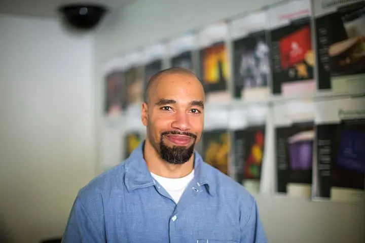 A man with a shaved head and goatee, wearing a blue shirt, stands indoors in front of a wall covered with various posters and images, smiling at the camera.