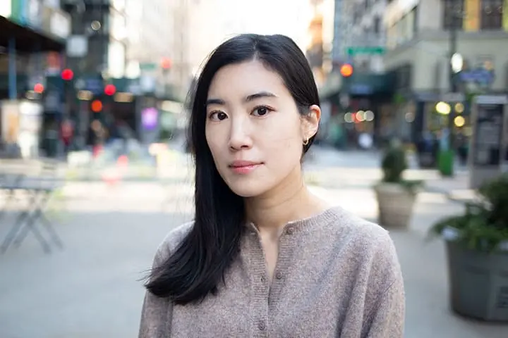 A woman with long black hair and a beige sweater stands outdoors in a city street, looking at the camera with a neutral expression. The background is blurred with buildings and street lights visible.