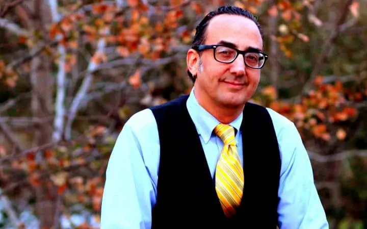 A man with short dark hair, glasses, and a slight smile wears a light blue shirt, yellow striped tie, and black vest, standing outdoors in front of autumn-colored trees.