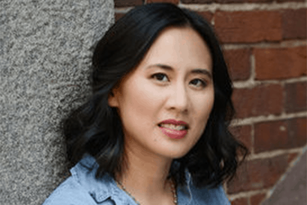 A woman with wavy dark hair and a light blue shirt stands in front of a brick wall, looking at the camera with a slight smile.