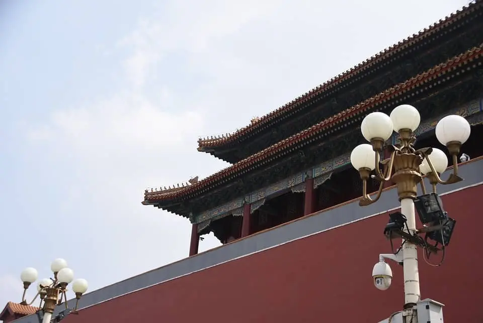 Traditional Chinese architecture with ornate, tiered roofs rises above a red wall; in the foreground, white globe street lamps evoke the global spirit of the PEN World Voices Festival under a partly cloudy sky.