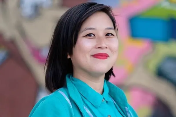 A woman with short dark hair and red lipstick smiles at the camera, wearing a teal shirt. The colorful, blurred background includes shades of pink, blue, yellow, and brown.