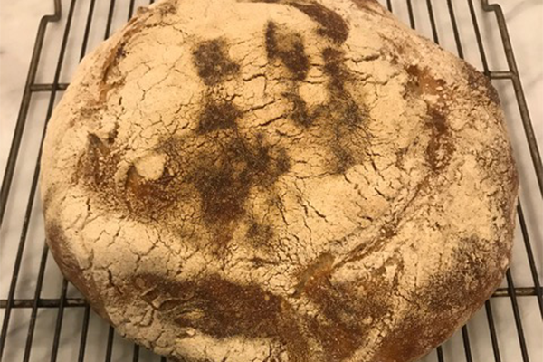 A round loaf of rustic bread with a golden-brown, cracked crust rests on a cooling rack. The surface is dusted with flour and has dark, baked spots.