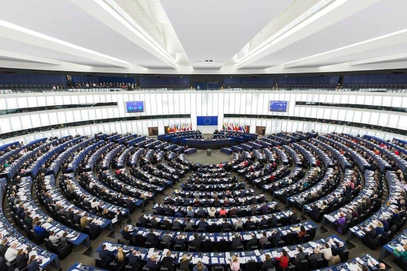 A wide-angle view of the European Parliament chamber filled with delegates seated in curved rows, facing a central podium with flags and large screens displaying information.