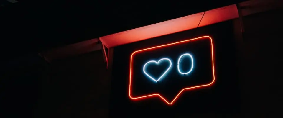 A neon sign shaped like a social media notification bubble displays a blue heart and the number zero, symbolizing zero likes, against a dark background.