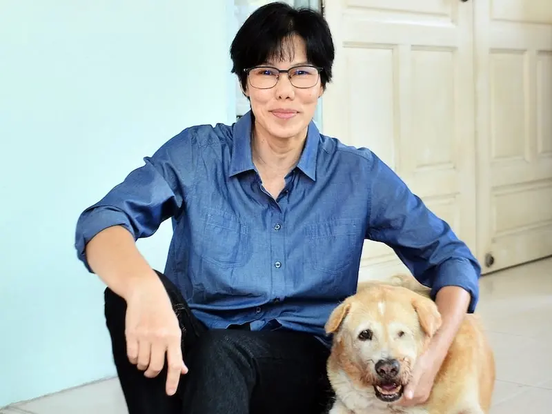 A person with short dark hair and glasses, wearing a blue shirt and black pants, sits on the floor indoors, smiling and gently holding a happy, light-colored dog beside them.