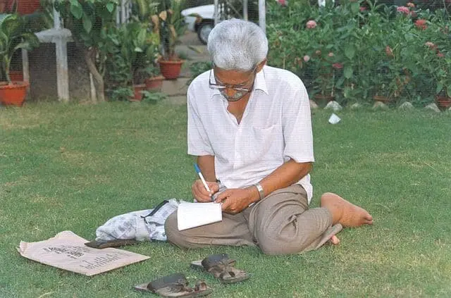 An elderly person with gray hair, resembling P. Varavara Rao, sits barefoot on grass, writing in a notebook. Sandals, a bag, and a paper bag lie nearby amid green plants and potted flowers in the background.