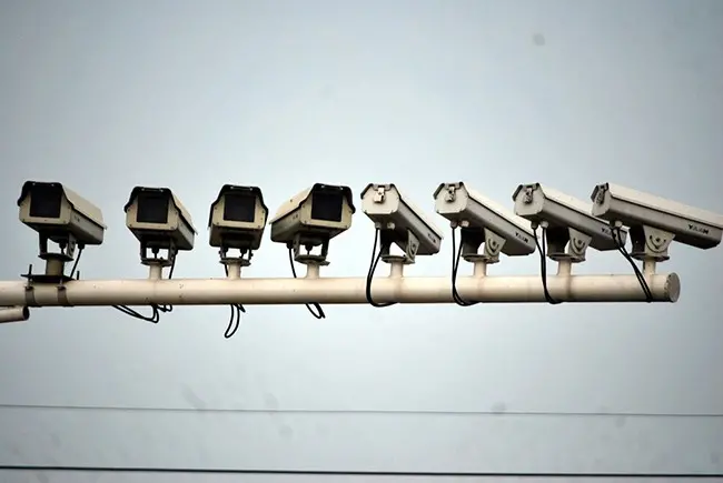 A row of eight surveillance cameras mounted on a horizontal pole against a plain sky background, with two electrical wires visible below.