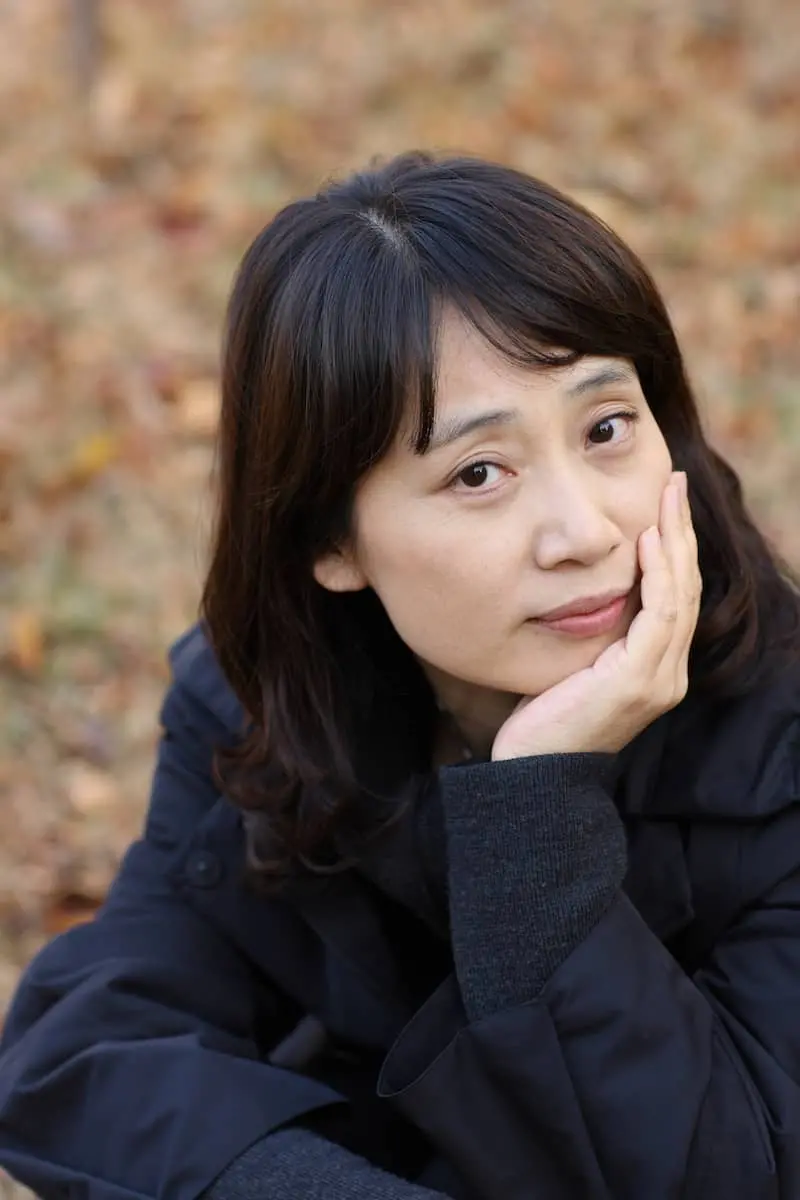 A woman with medium-length dark hair wearing a dark coat rests her face on her hand and looks thoughtfully at the camera. The background is out of focus with autumn leaves on the ground.