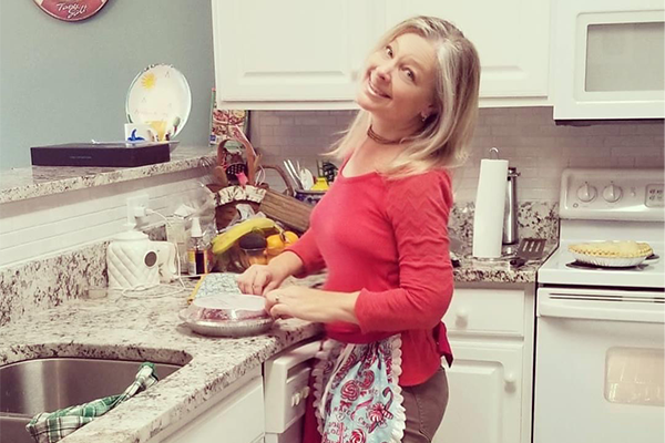 A woman smiling in a kitchen while wrapping food with plastic wrap on a counter beside a sink. Fruits, kitchen items, and a pie on the stove are visible in the background.