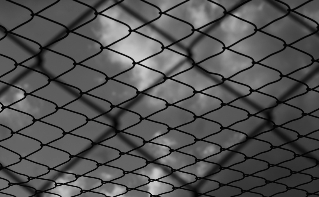 A close-up view of a chain-link fence in black and white, with a cloudy sky visible in the background. The fence is in sharp focus, while the clouds are slightly blurred.