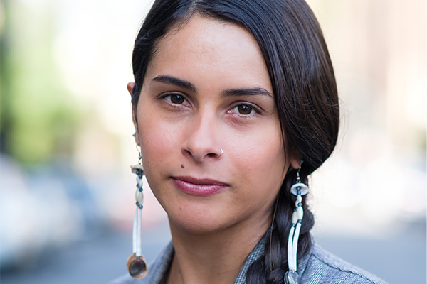 A woman with long dark hair in a side braid, wearing dangling earrings and a nose ring, looks confidently at the camera with a neutral expression. The background is blurred outdoor scenery.