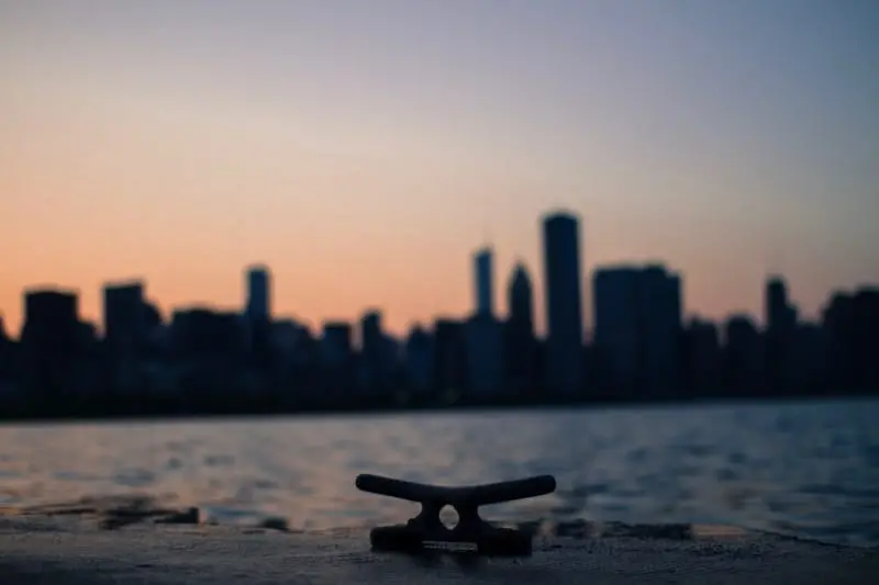 A boat cleat sits in the foreground with a city skyline and calm water in the background at sunset, creating a silhouette effect against the colorful sky.