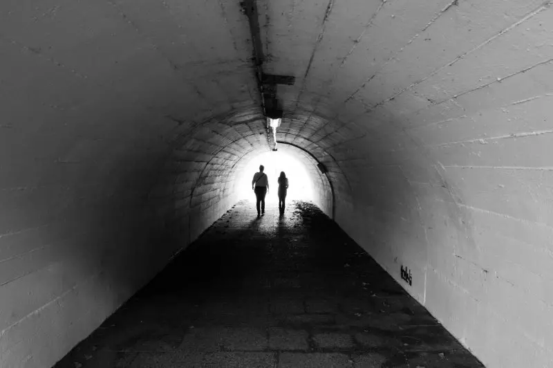 Two people walk side by side through a concrete tunnel toward bright daylight at the tunnel’s end, creating strong silhouettes against the illuminated exit.