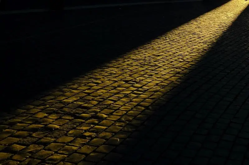 A narrow beam of golden sunlight illuminates a path of cobblestone pavement, creating a bright streak across the otherwise dark, shadowy ground.