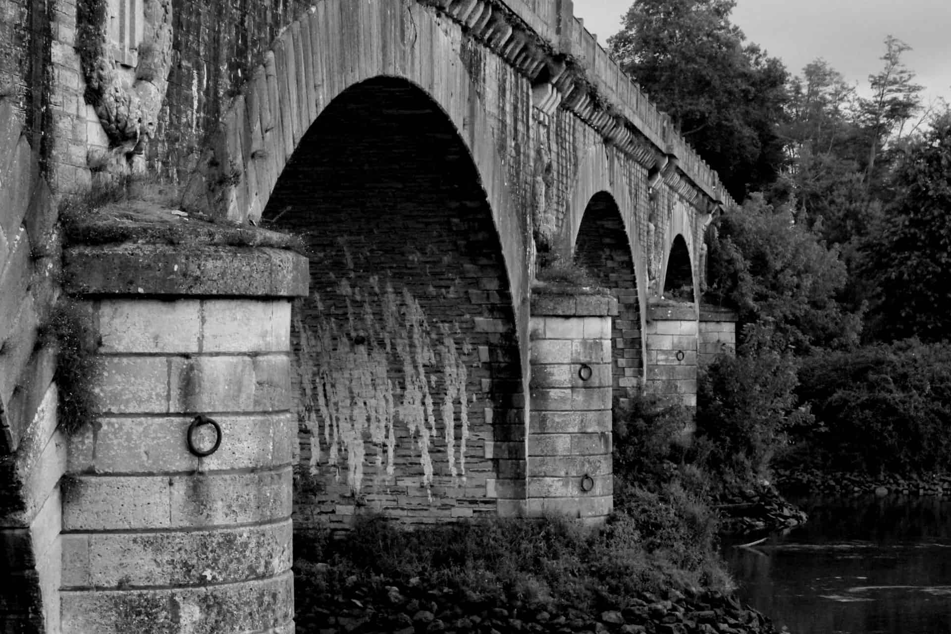 A black and white photo of an old stone bridge with multiple arches crossing over a river, surrounded by trees and vegetation. The weathered stonework and water beneath create a historic, serene atmosphere.