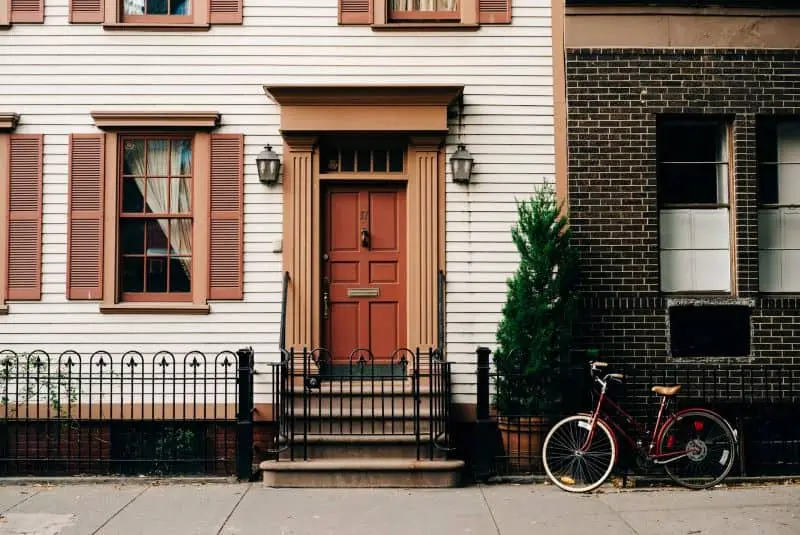 A red door with decorative trim and two lanterns is centered between shuttered windows on a white and brown townhouse. A black iron fence and a red bicycle are in front, and greenery grows by the steps.