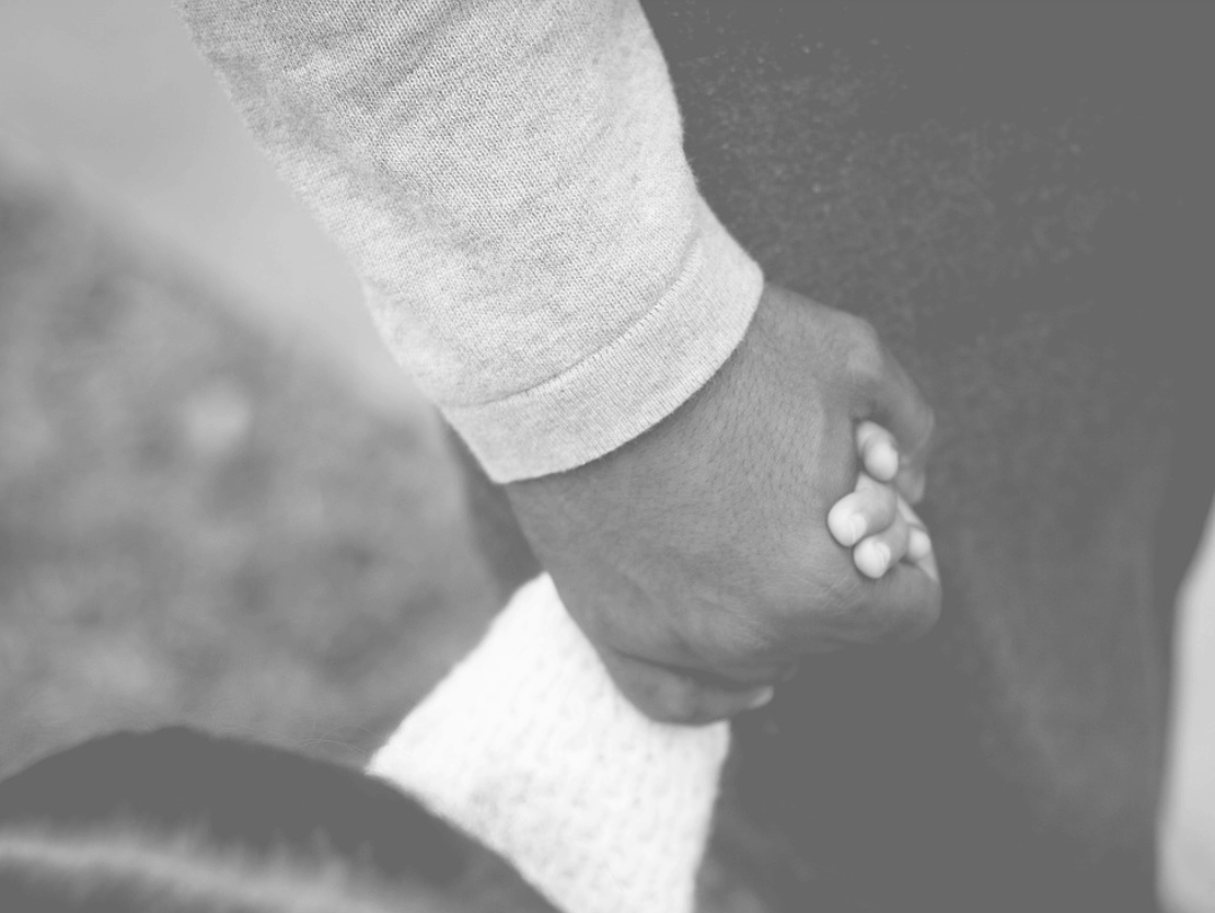 A close-up, black-and-white image shows an adult and a child holding hands. The adults hand gently envelops the smaller hand, suggesting care and protection.