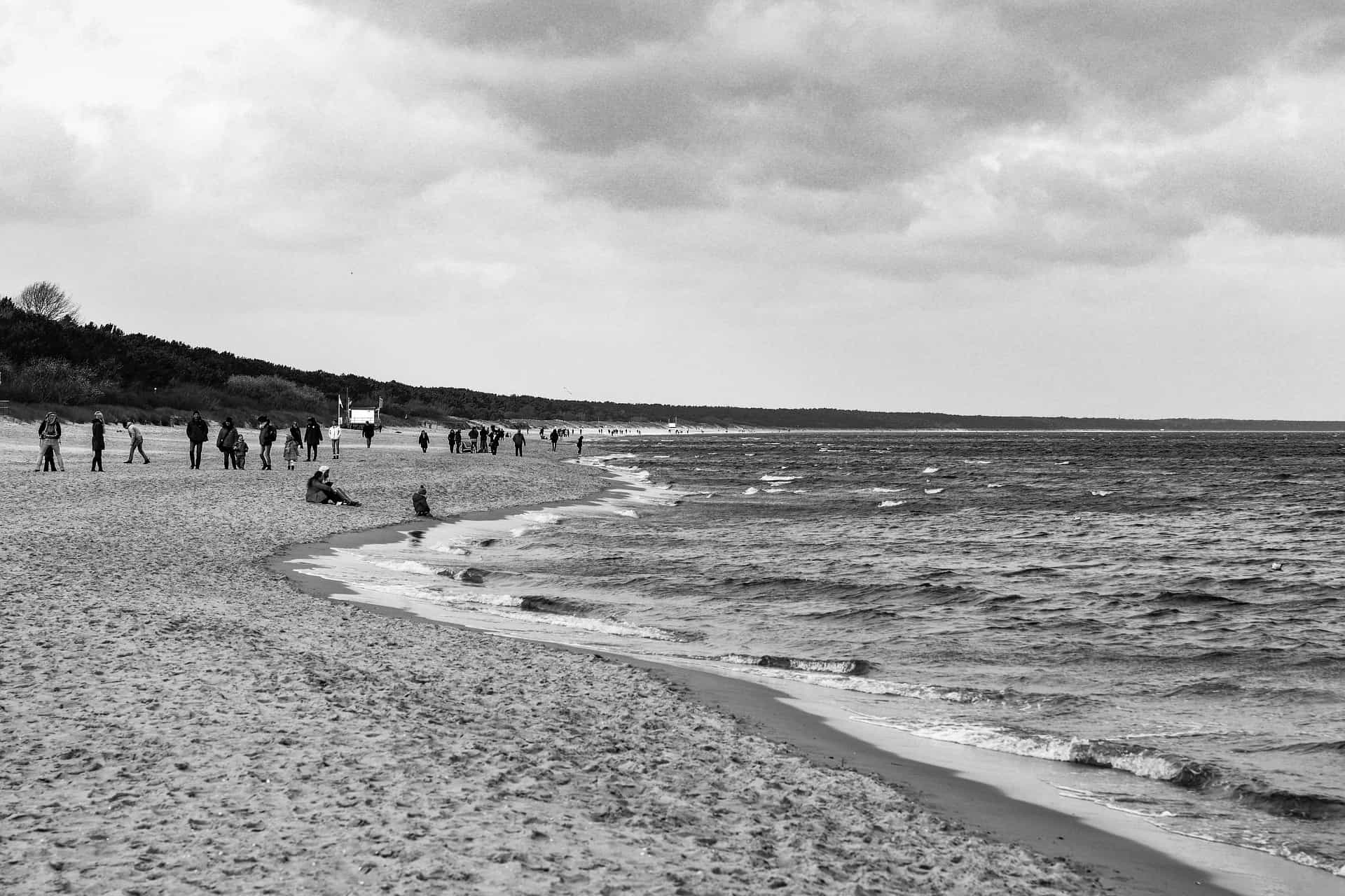 Black and white photo of a beach with gentle waves; people are walking along the shoreline, some standing and others sitting, with trees in the background and a cloudy sky overhead.