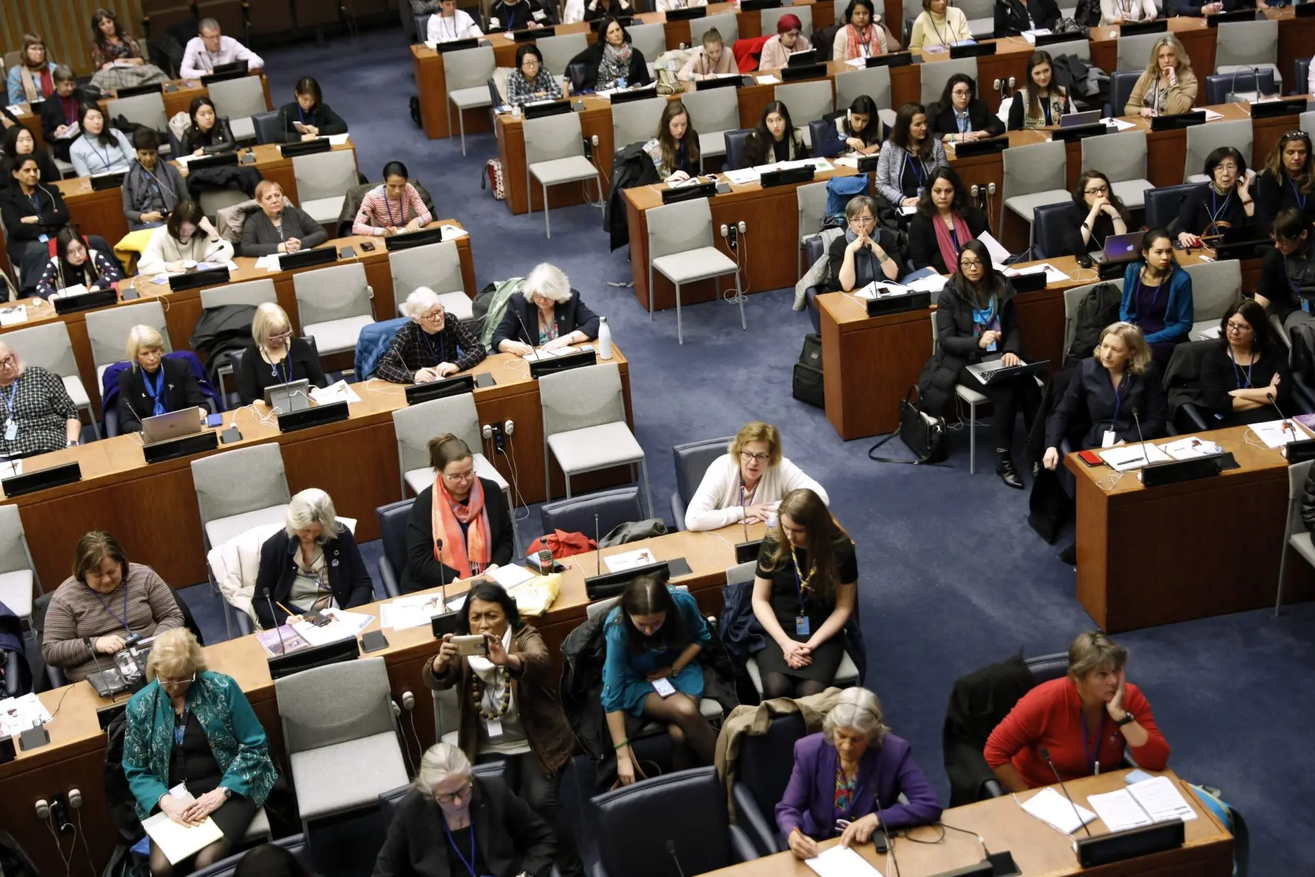 A large group of people, mostly women, sit and listen attentively in a formal auditorium or conference room with rows of desks, notebooks, and microphones.