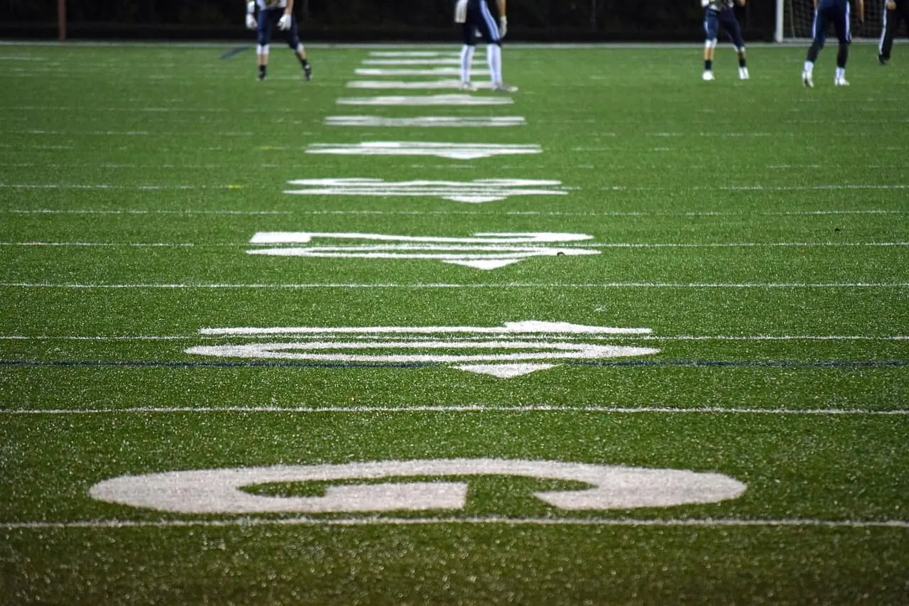 Close-up view of an American football field, focused on the yard lines and numbers, with a few players standing and walking in the blurred background.