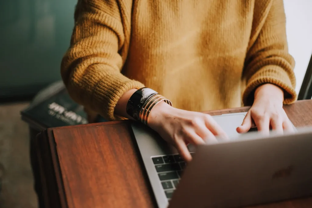 A person wearing a mustard yellow sweater and bracelets is typing on a laptop at a wooden desk, perhaps drafting a Book Ban Open Letter. Only their arms and hands are visible. A closed book is partially seen nearby.