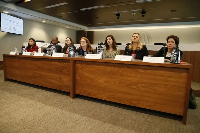 Seven people sit behind a long wooden table with microphones and nameplates, attending a formal meeting or panel discussion in a modern conference room.