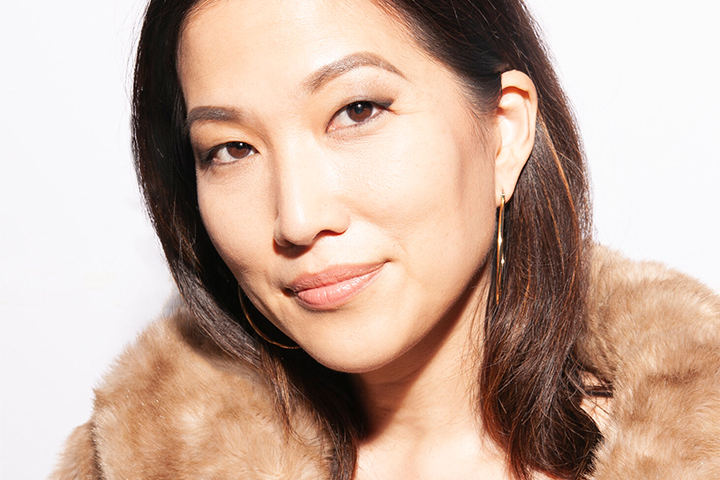 A woman with straight dark hair, wearing hoop earrings and a tan fur coat, looks confidently at the camera against a plain white background.