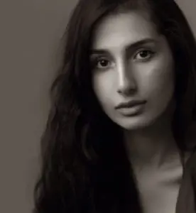 Black and white portrait of a woman with long, wavy hair. She is looking directly at the camera with a calm and neutral expression. The background is plain and soft-focused.