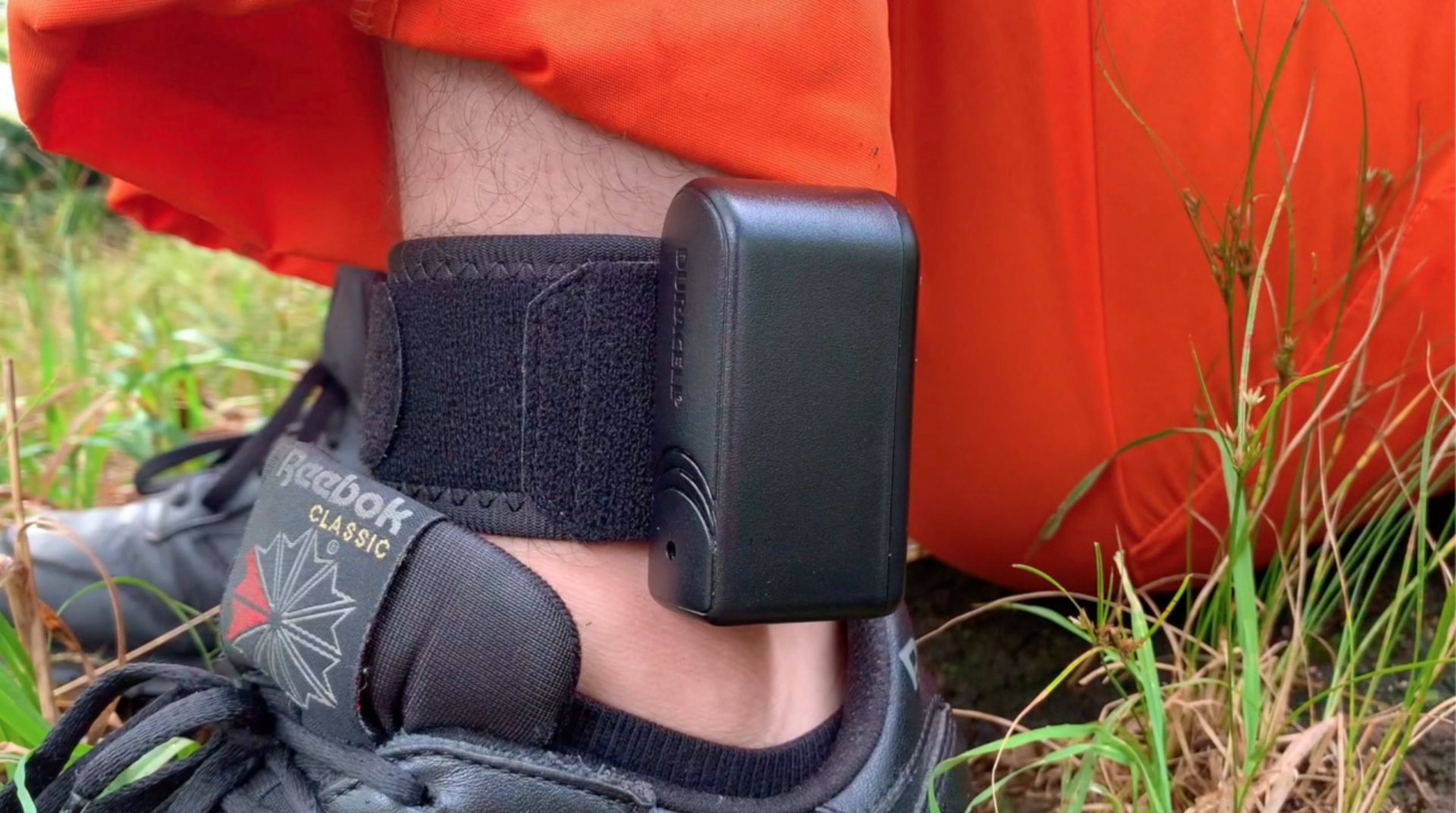 A close-up of a persons ankle wearing a black electronic monitoring ankle bracelet, with orange pants and a black Reebok sneaker, outdoors on grass.