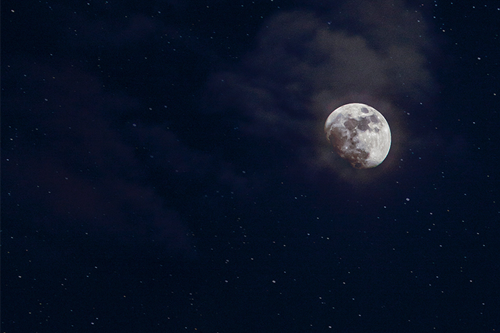 A bright, partially cloud-covered moon shines in a dark, star-filled night sky.
