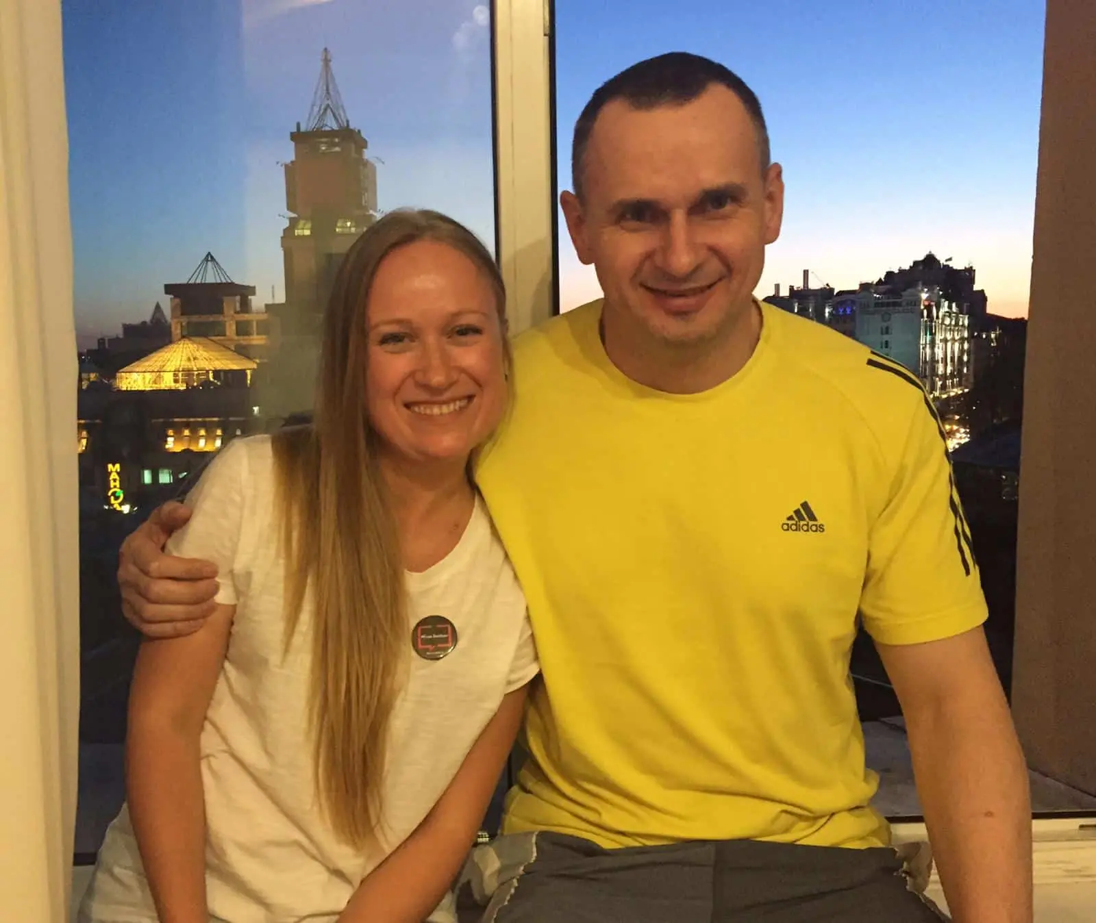 A smiling woman and man sit close together by a window at sunset, with city buildings in the background. The woman has long blonde hair and wears a white t-shirt; the man wears a yellow Adidas shirt.