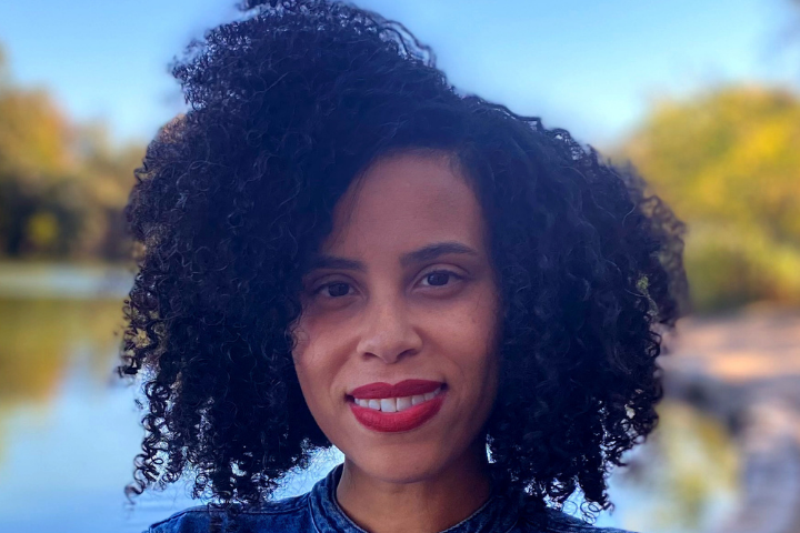 A woman with curly black hair and red lipstick smiles slightly while standing outdoors near a body of water, with trees and a blue sky in the background.