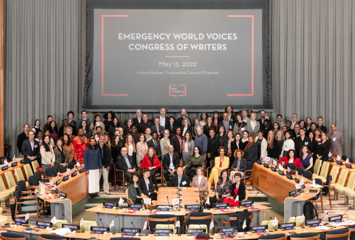 A large group of people poses for a photo in a conference room with desks and microphones. A screen behind them reads Emergency World Voices Congress of Writers, May 13, 2022, United Nations Trusteeship Council Chamber.