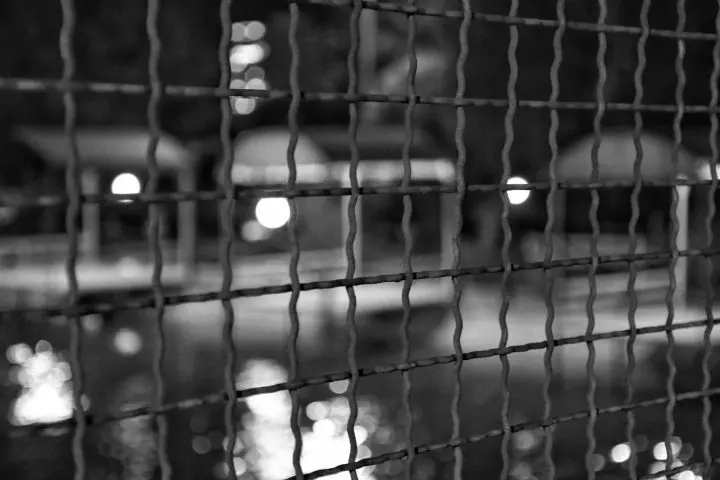 A close-up of a wire fence in sharp focus with blurred lights and structures in the background, creating a bokeh effect in a nighttime black-and-white scene.
