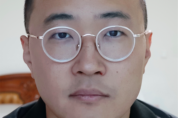 A person with short hair wearing round, clear-framed glasses looks directly at the camera with a neutral expression. The background is softly blurred, featuring part of a wooden chair.